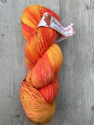 Ein handgefärbter Strang 6-fach Sockenwolle in intensiven Gelb-, Orange- und Pinktönen. Der Strang liegt auf hellem Holz; das Etikett zeigt das Herzfasern-Logo in Pink-Orange.